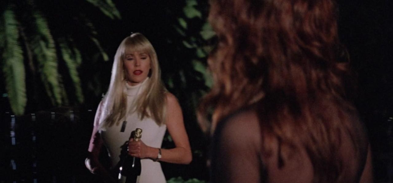 Eine blonde Frau in weißem Kleid hält eine Champagnerflasche, während eine andere Frau mit roten Haaren im Vordergrund steht.