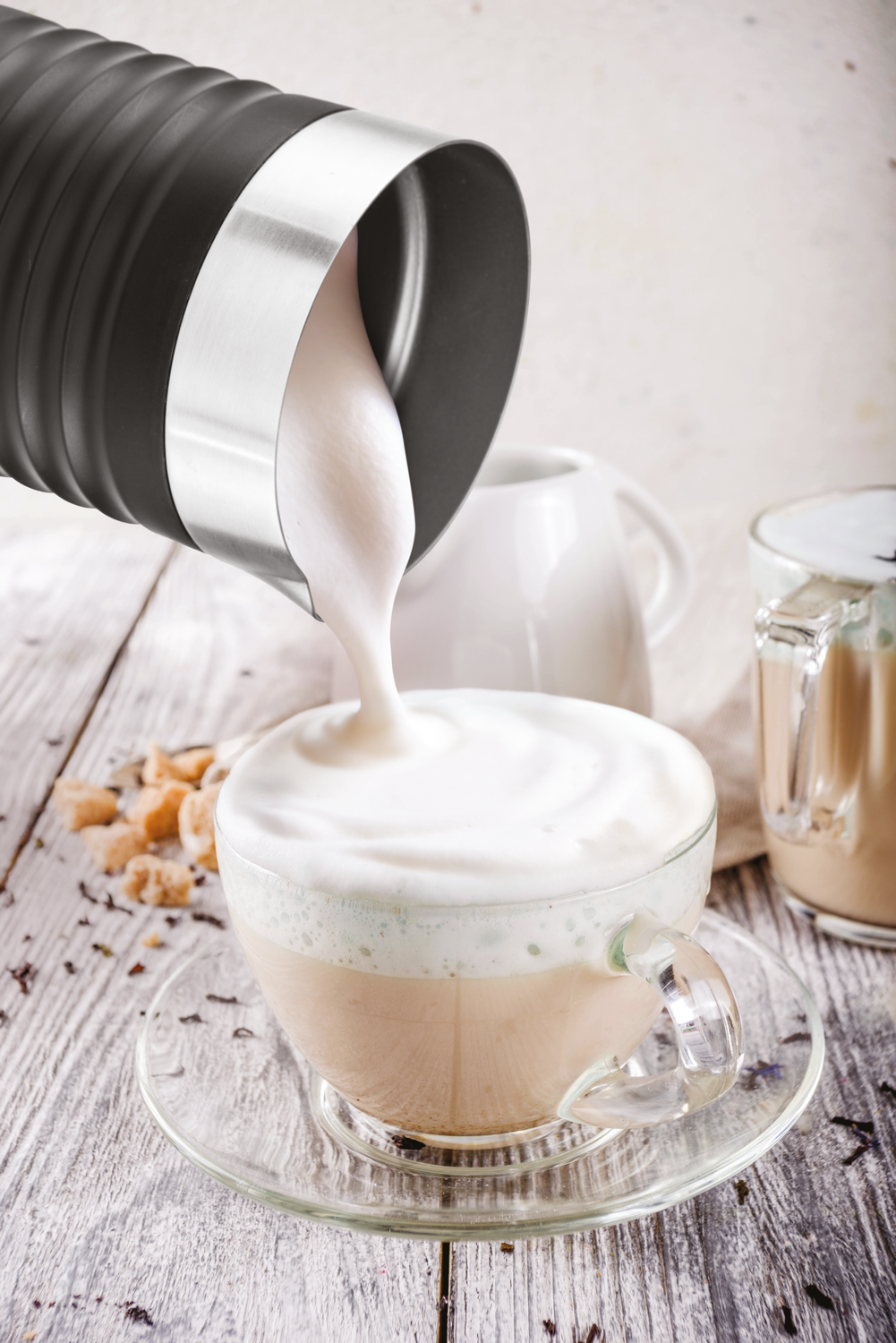 Un espumador negro vierte espuma cremosa en una taza de café de vidrio sobre una mesa de madera.