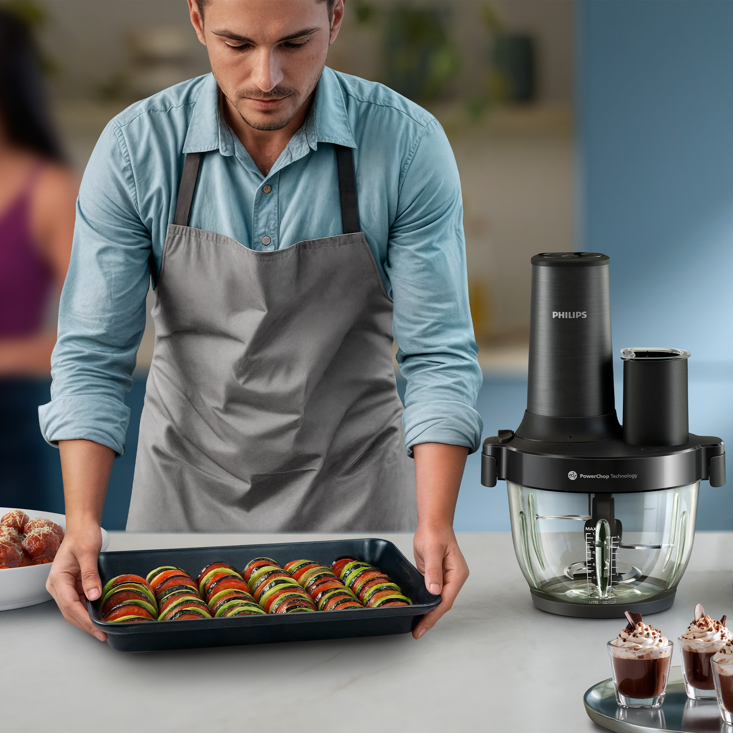 Man in apron holds baking dish with layered vegetables next to Philips food processor.