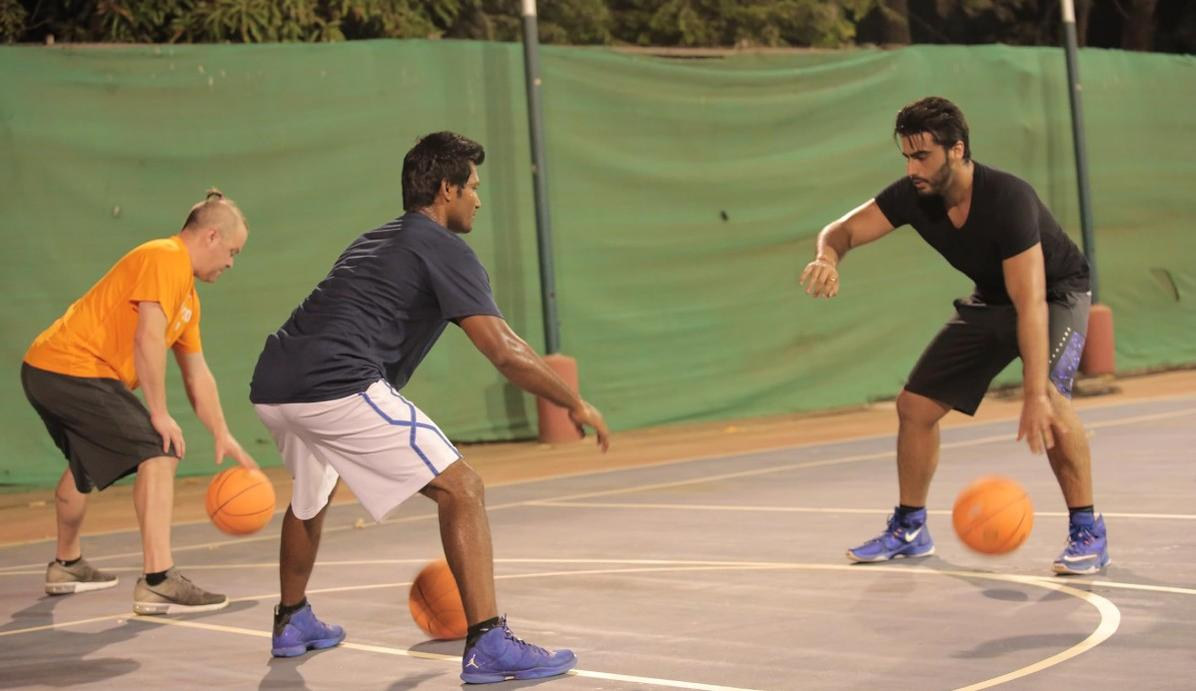 Drei Männer spielen Basketball auf einem Freiplatz mit grünen Zäunen im Hintergrund.