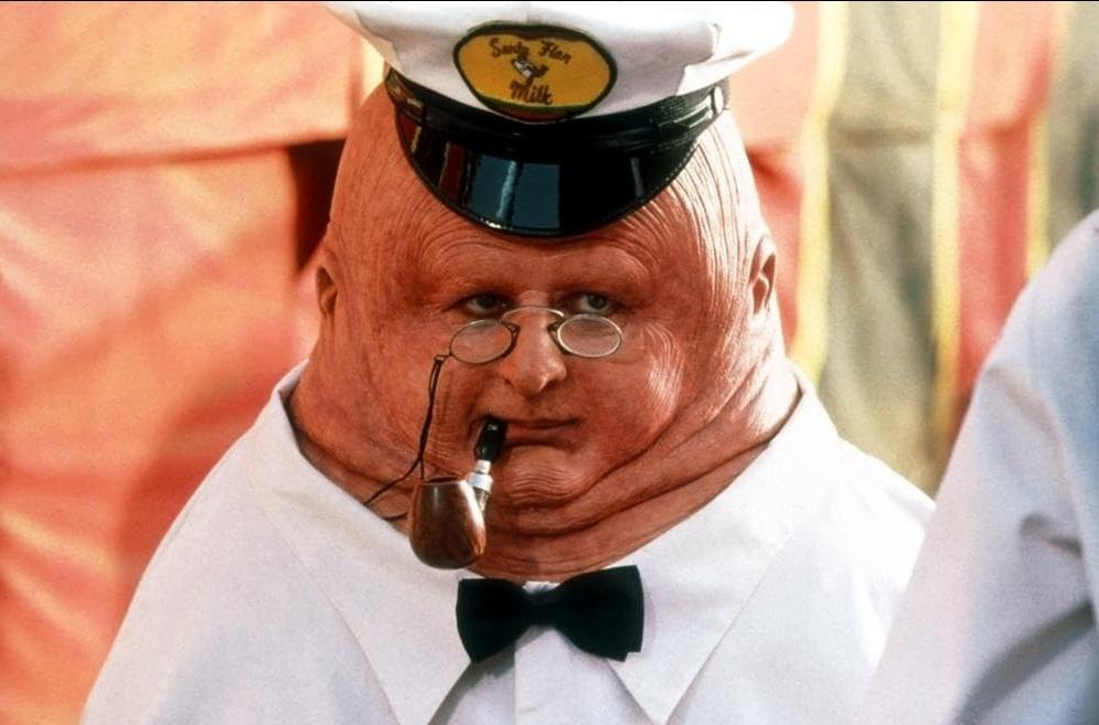 Ein Mann mit zerknitterter Haut, Brille und Pfeife trägt eine weiße Uniform mit Fliege.