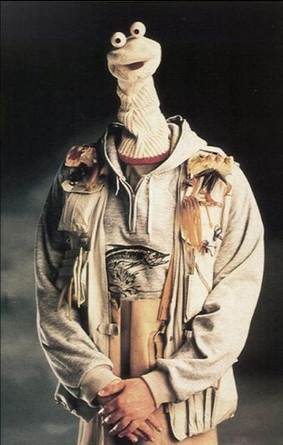 Ein Sockenpuppen-Charakter trägt eine Kapuzenjacke mit Fischmotiv und eine Anglerweste.