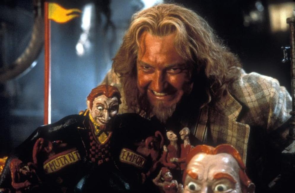 Ein Mann mit langen Haaren und Bart lächelt, umgeben von Puppen und einem gelben Banner.