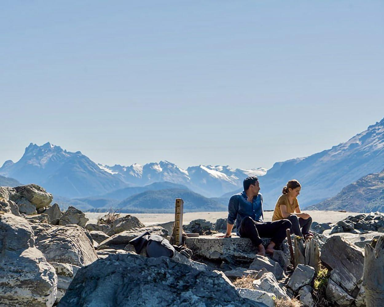 Zwei Personen sitzen auf Felsen mit schneebedeckten Bergen im Hintergrund.