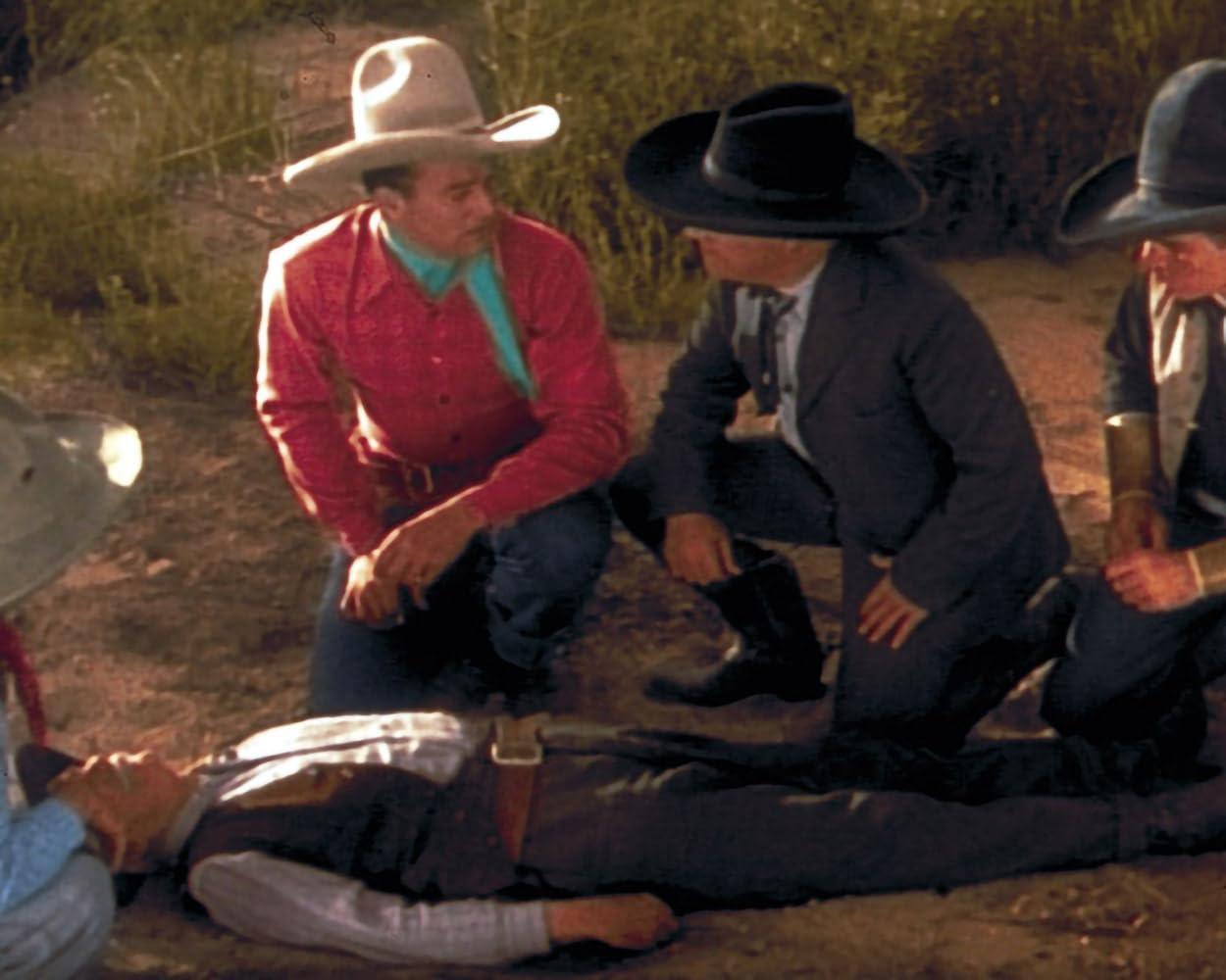 Männer in Cowboykleidung knien um einen liegenden Mann auf staubigem Boden.