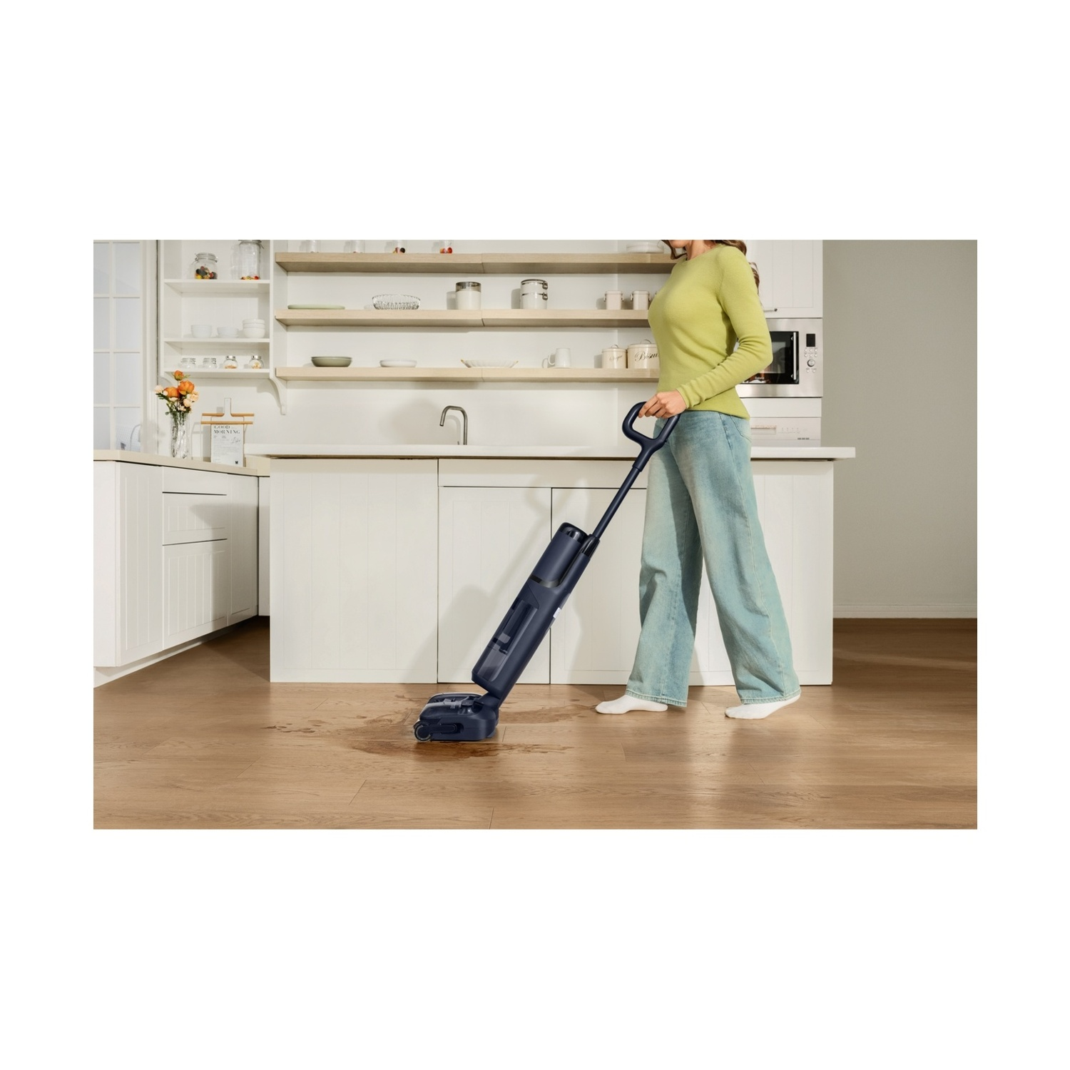 Una donna pulisce un pavimento di legno in cucina con un aspirapolvere blu.