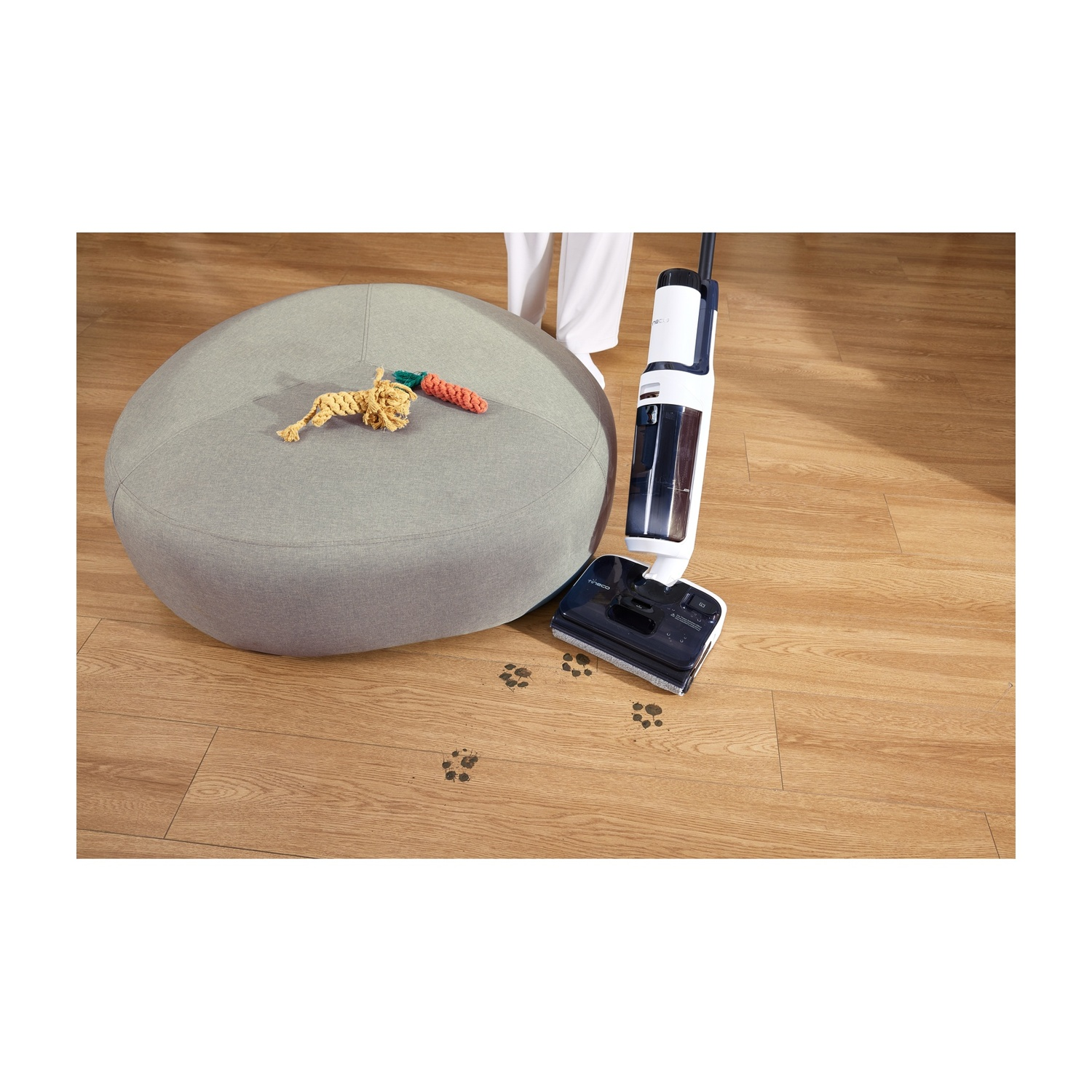 Un aspirapolvere lavapavimenti accanto a un pouf grigio con giocattoli. Impronte di zampe sul pavimento.