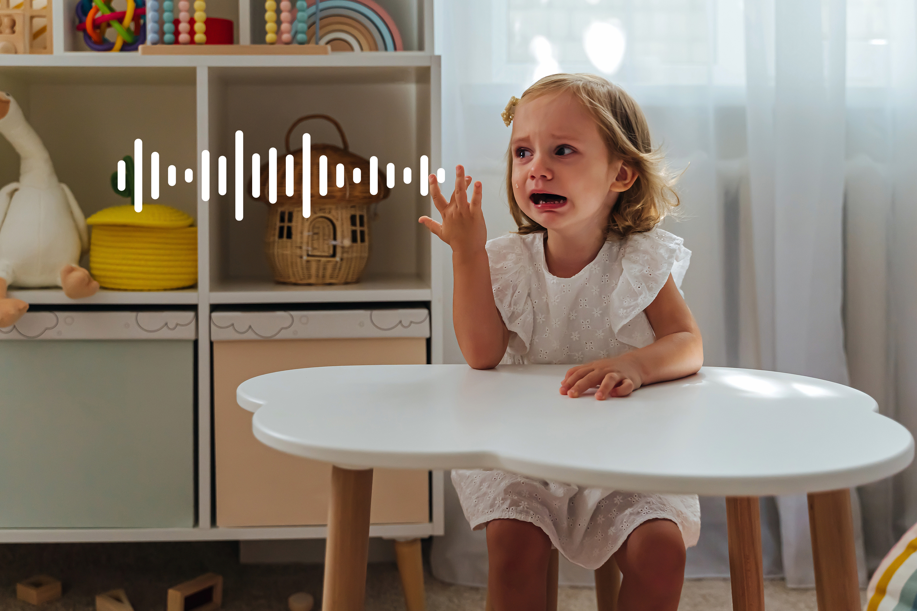 Ein weinendes kleines Mädchen sitzt an einem Tisch im Kinderzimmer. Spielzeug ist im Regal.