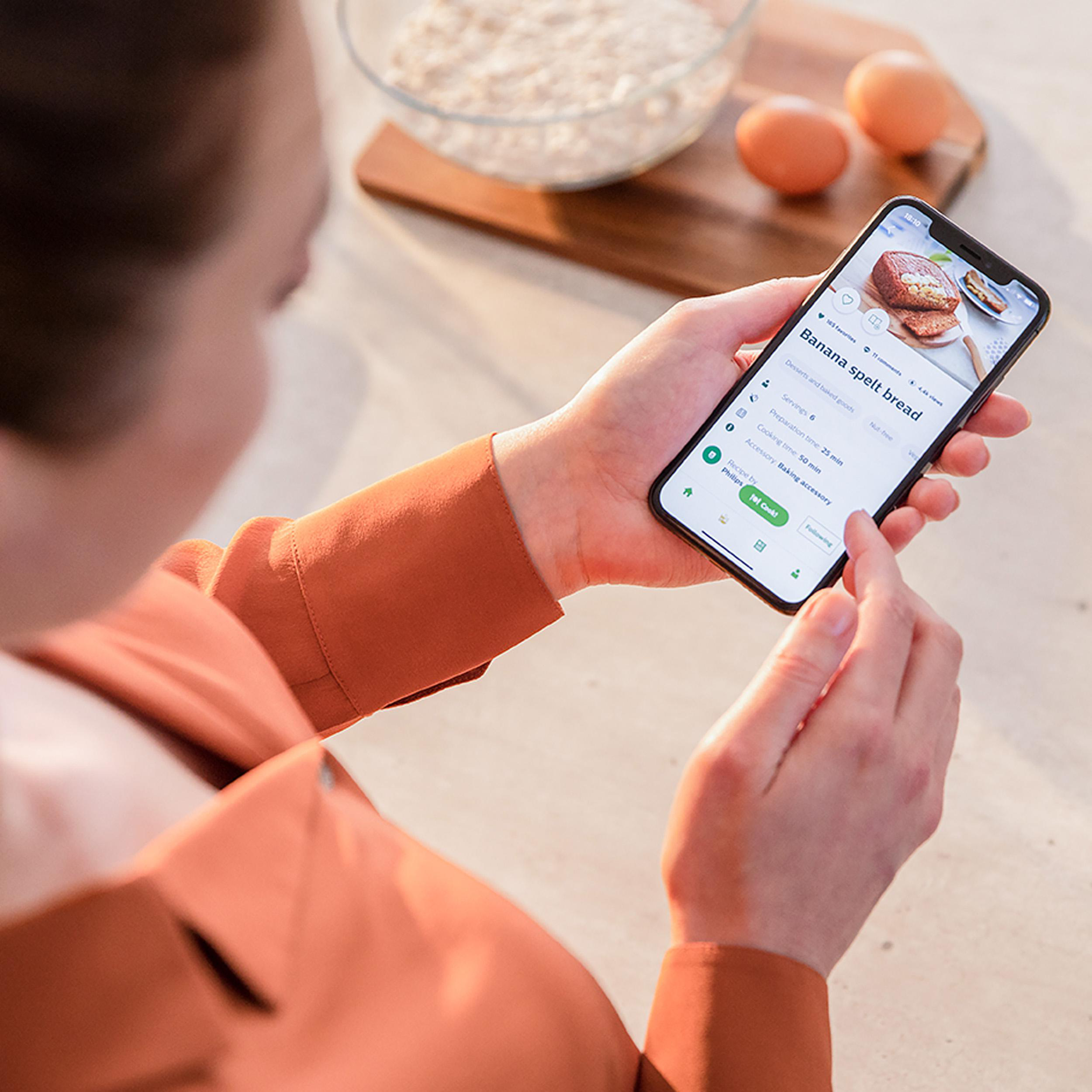 Eine Person hält ein Smartphone mit einem Rezept für Bananen-Dinkelbrot. Im Hintergrund Eier und Schüssel.