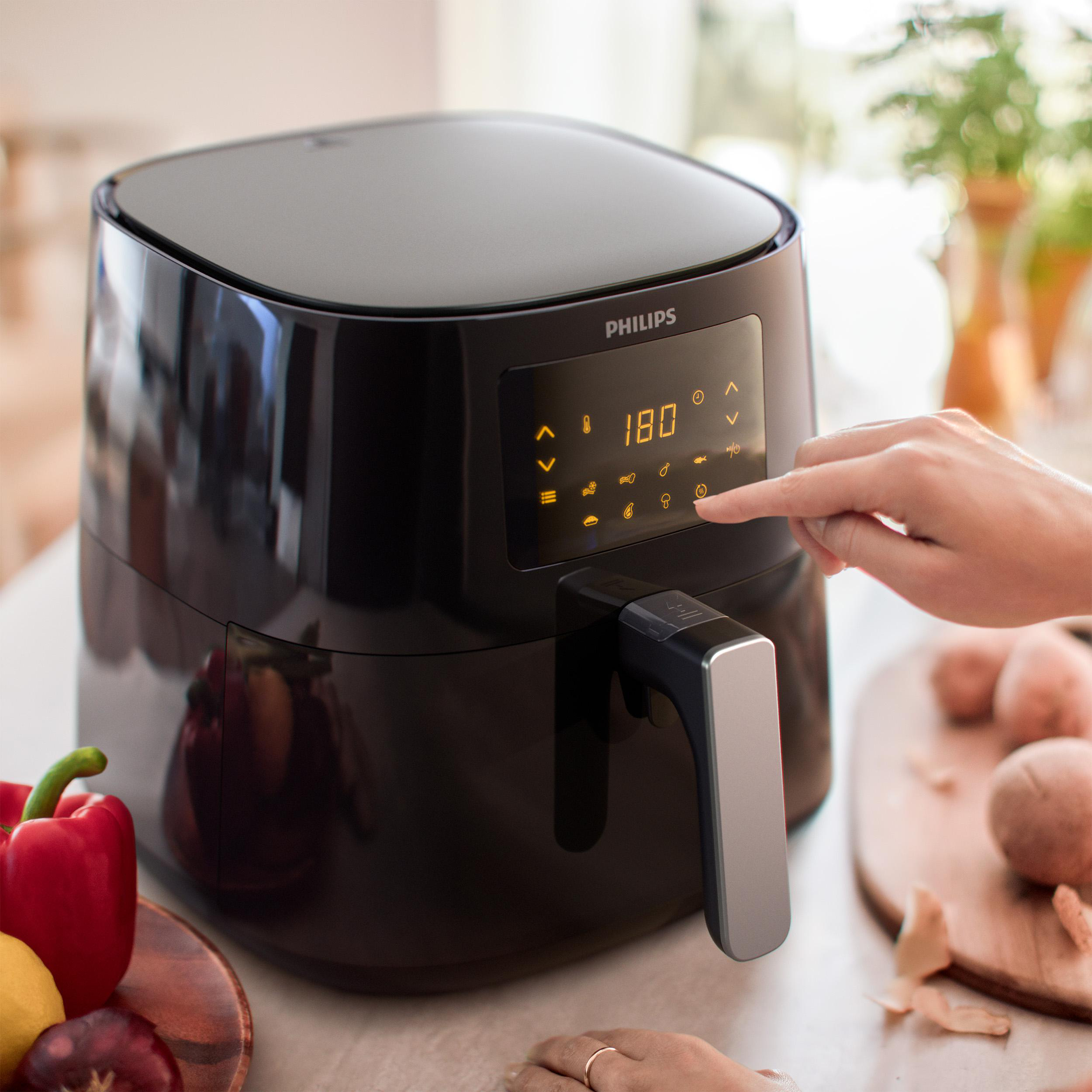 Eine Person bedient eine schwarze Heißluftfritteuse mit digitaler Anzeige. Zwiebeln, Paprika und Zitrone liegen daneben.