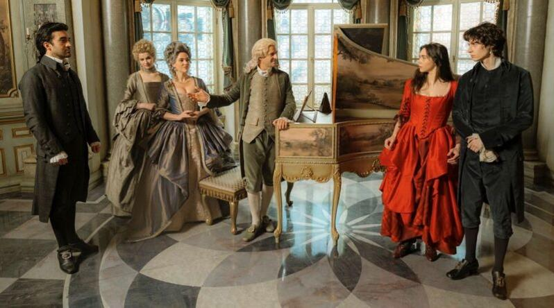 Sechs Personen in historischer Kleidung stehen in einem opulenten Raum vor einem Cembalo.