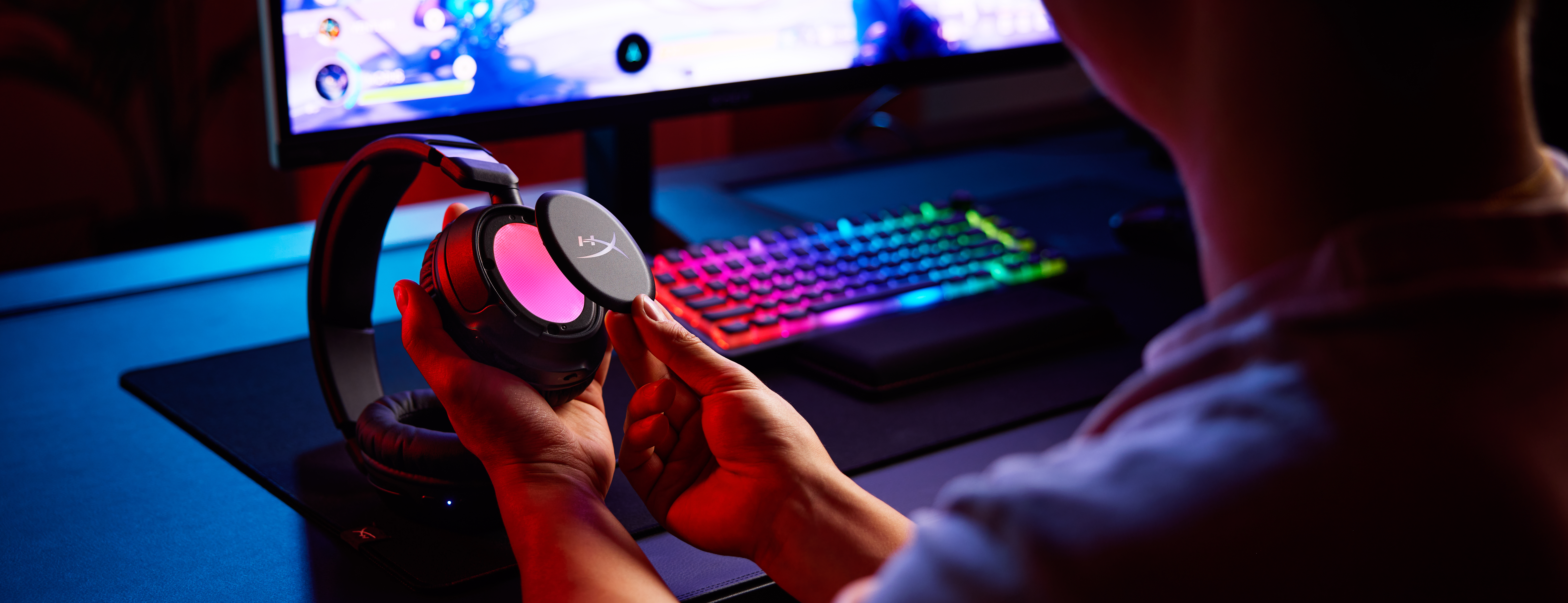 Una persona sostiene auriculares con iluminación rosa frente a un teclado y monitor iluminados.