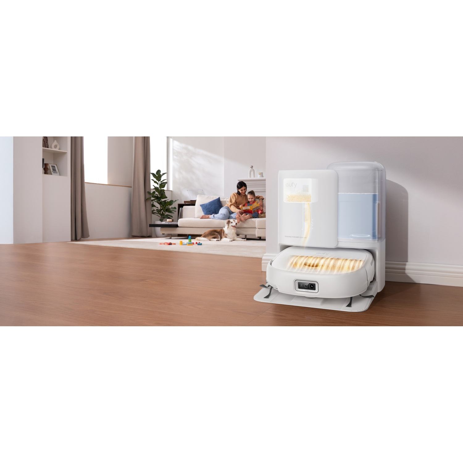 Un robot aspirateur blanc avec station de charge sur un sol en bois. Une femme et un enfant sont assis sur le canapé au fond.