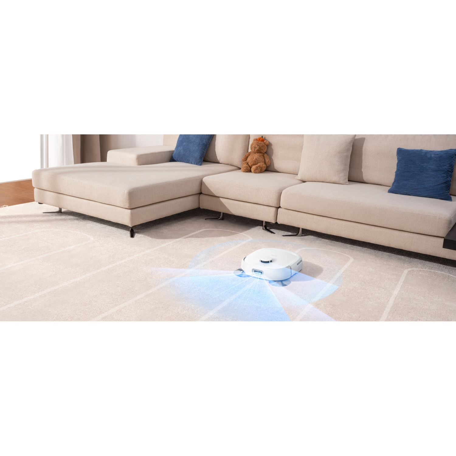 Un robot aspirateur blanc nettoie un tapis devant un canapé beige avec un ours en peluche.