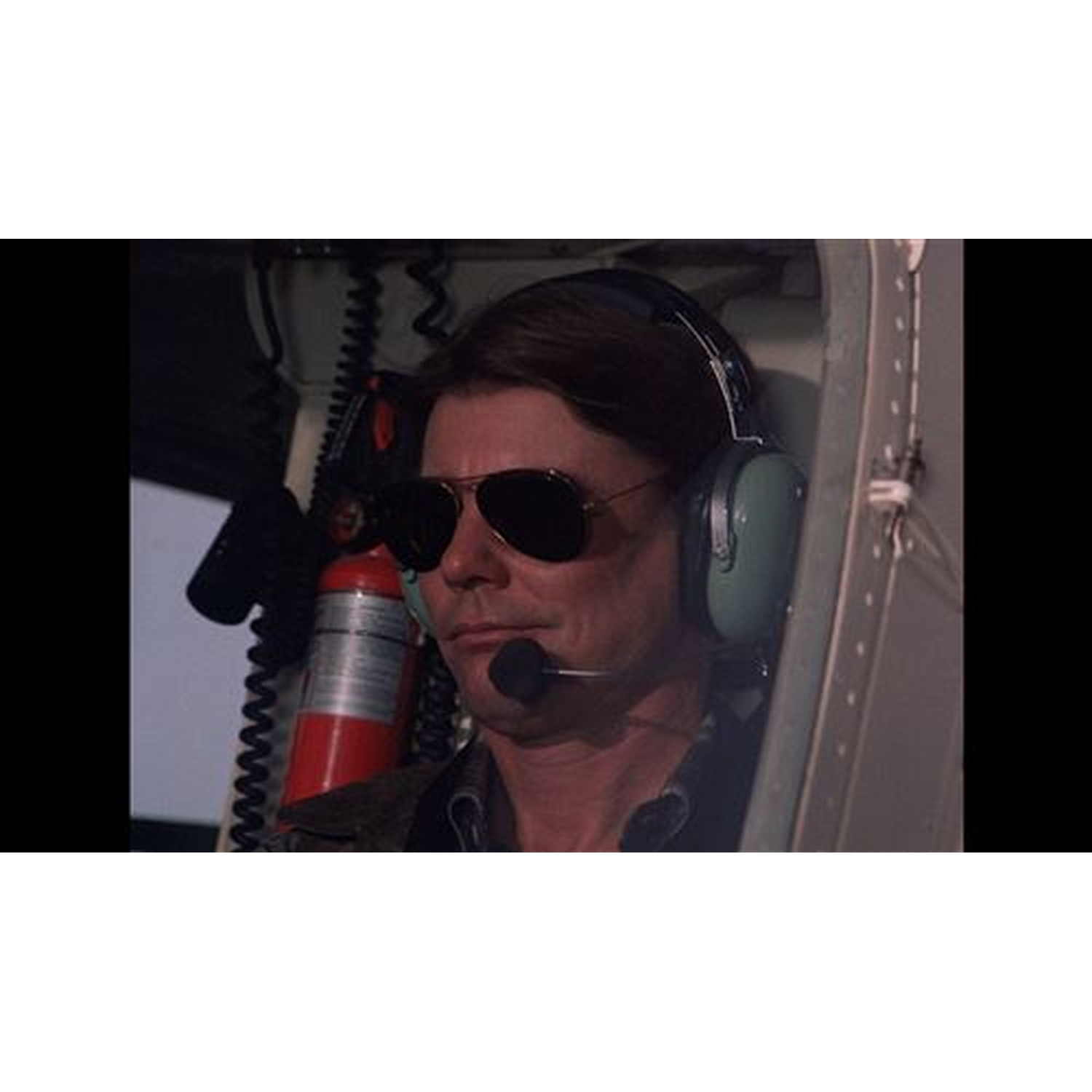 Un homme en lunettes de soleil et casque avec microphone est assis dans un véhicule. Un extincteur rouge est visible derrière lui.
