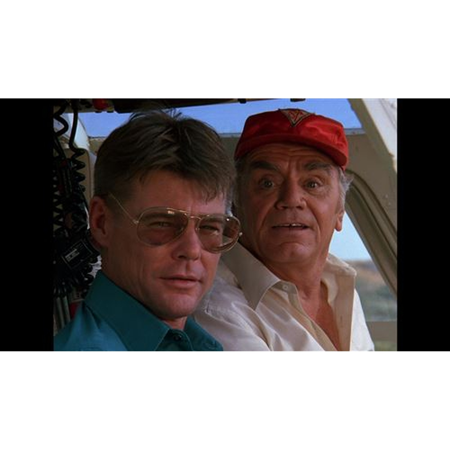 Deux hommes, l'un en chemise turquoise et lunettes d'aviateur, l'autre en casquette rouge et chemise blanche, regardent devant eux.