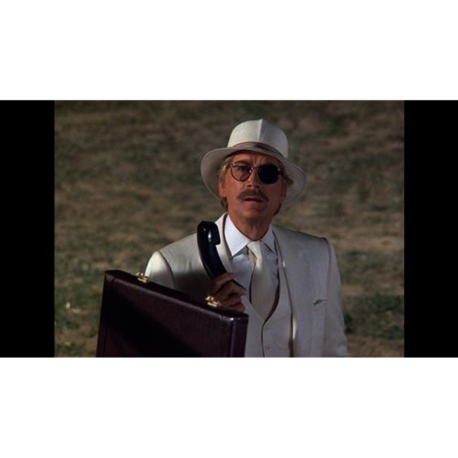Un homme en costume blanc, chapeau et cache-œil tient un combiné téléphonique noir et une mallette, regardant devant lui.