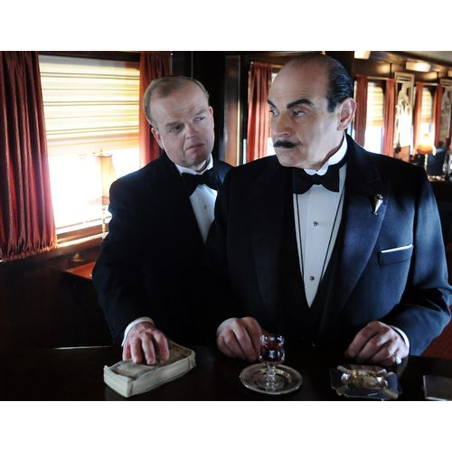 Deux hommes en smoking. L'un pose de l'argent sur une table, avec un verre et un cendrier.