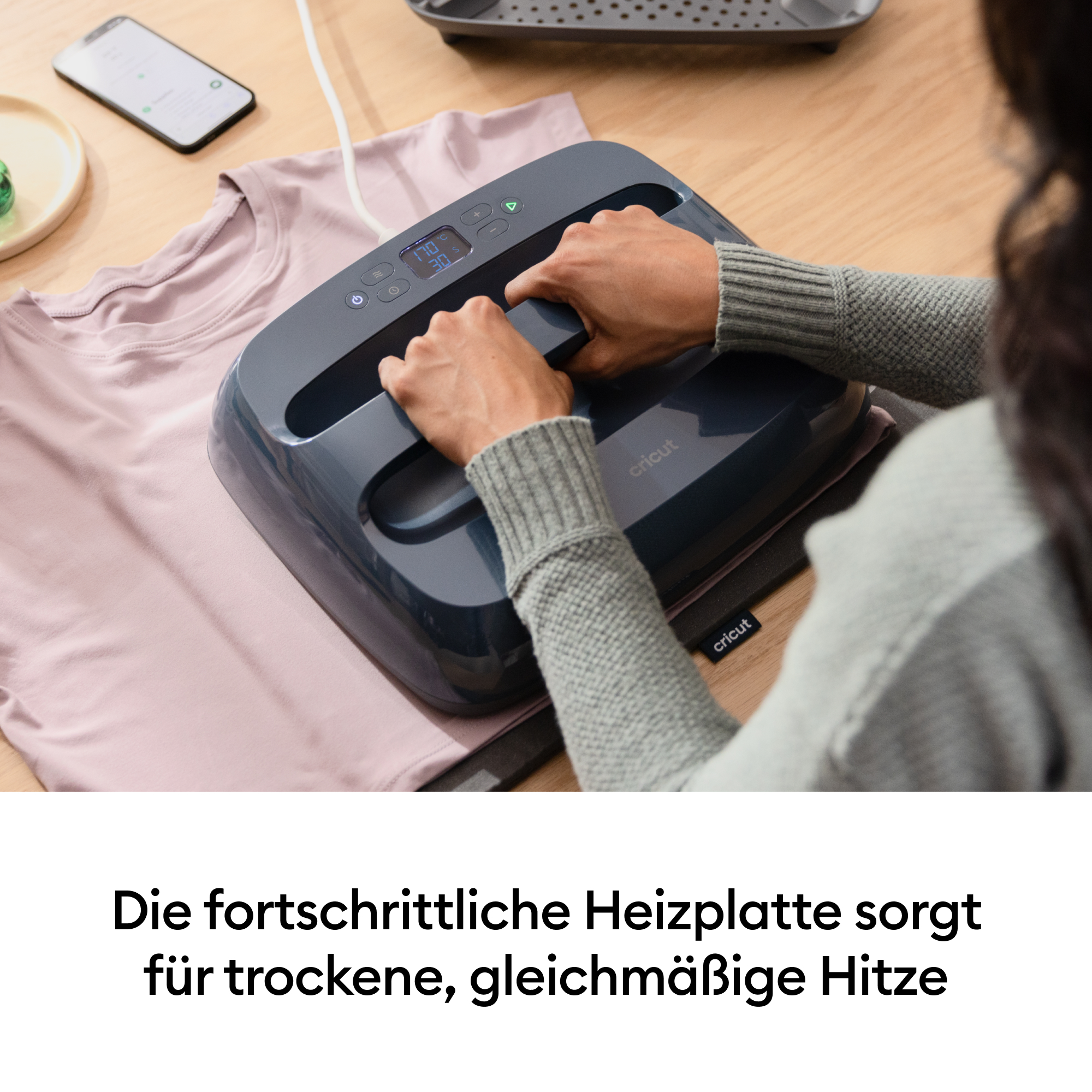 Eine Person nutzt eine Heißpresse auf einem rosa T-Shirt. Ein Handy liegt daneben.