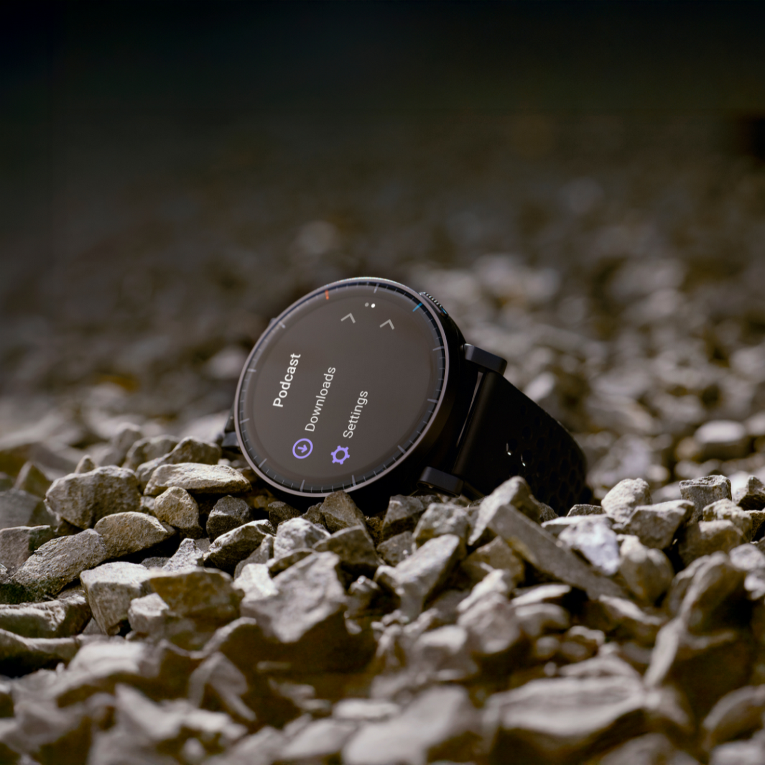 Un reloj inteligente negro descansa sobre piedras grises. La pantalla muestra el menú.