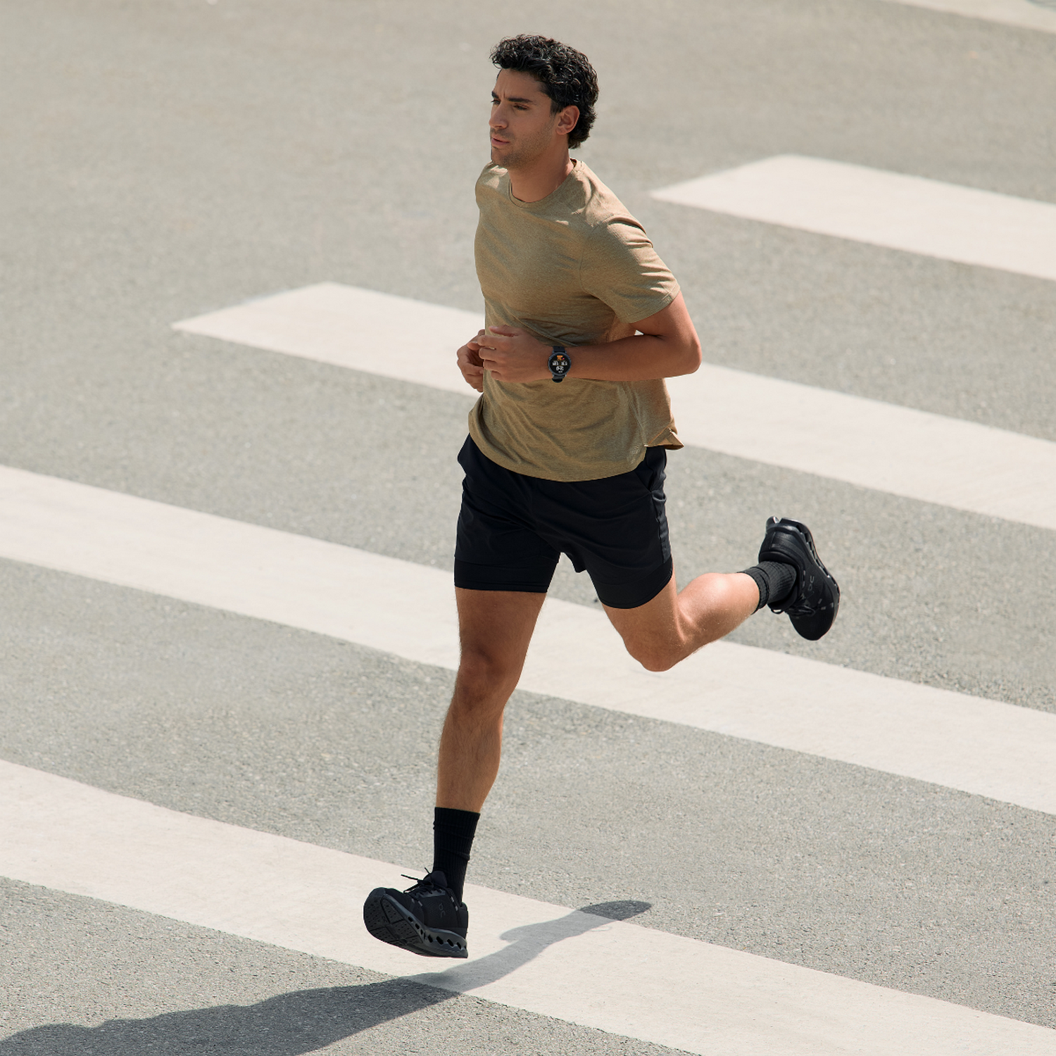 Un hombre con camiseta beige y pantalones cortos negros corre por un paso de cebra, su cuerpo en movimiento con un pie levantado.