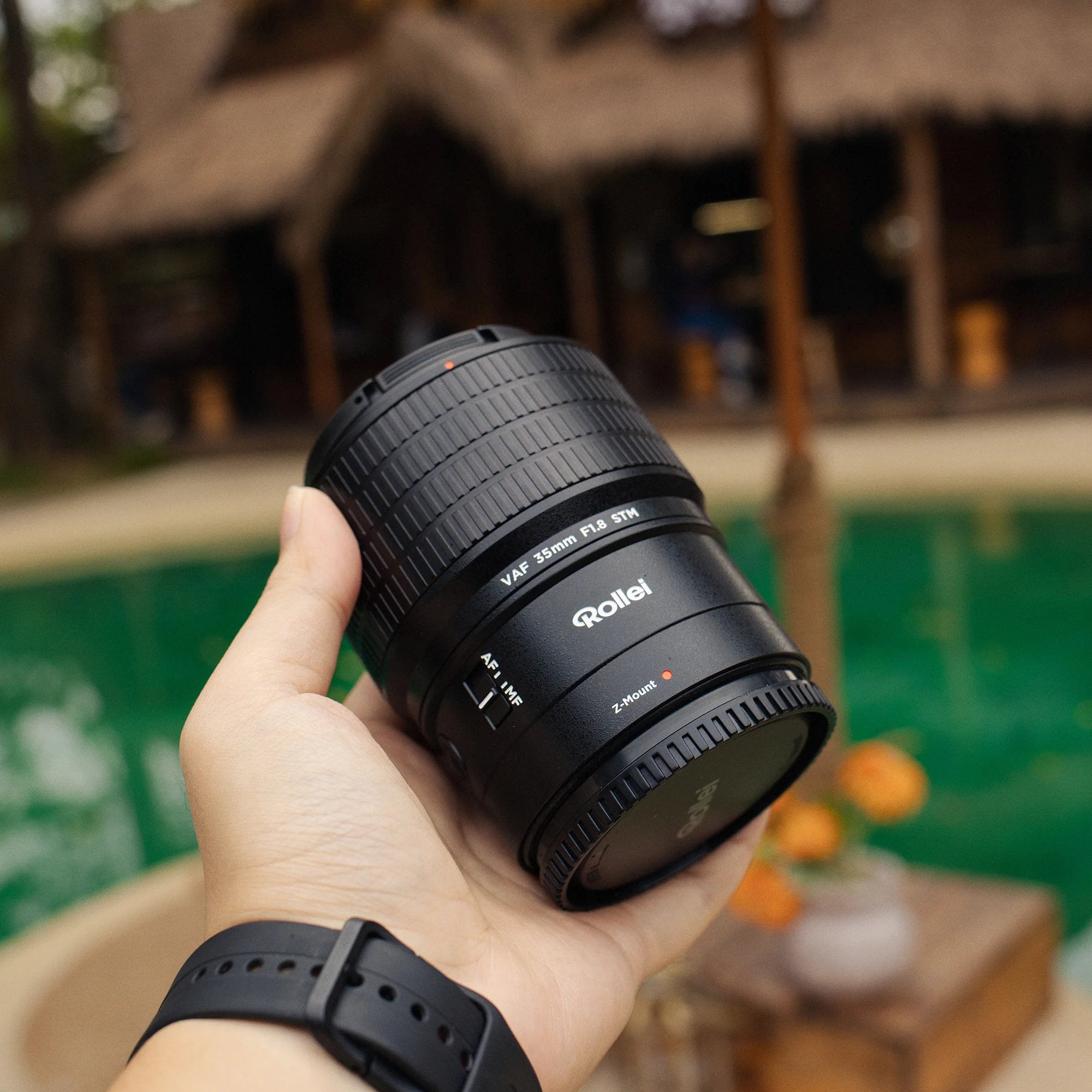 Eine Hand hält eine Rollei Kameraobjektiv vor einem grünen Pool und Hütten.