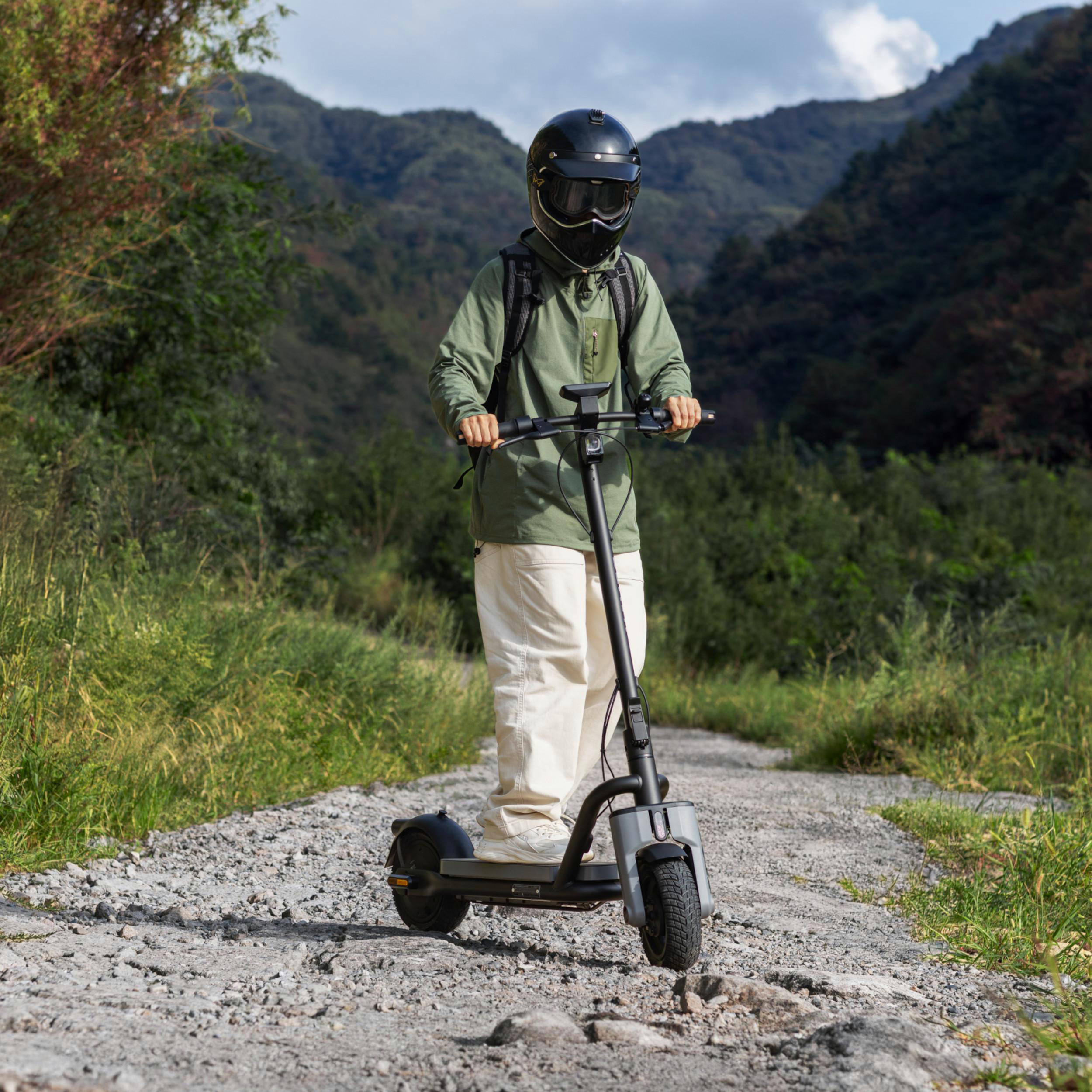 Person mit Helm auf Roller auf Schotterweg in bergiger Landschaft.