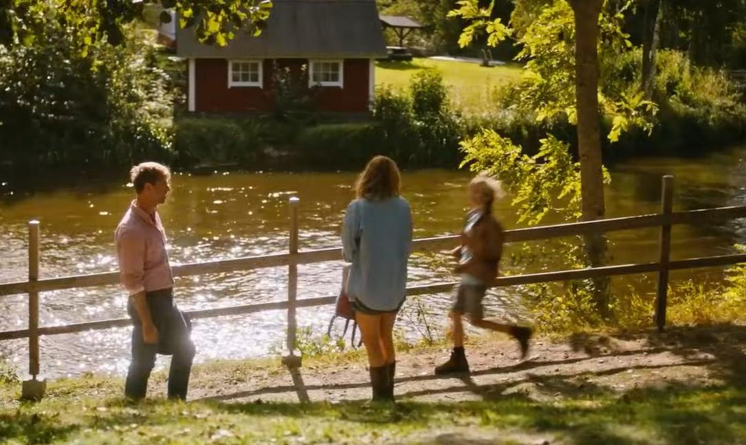 Drei Personen am Flussufer mit einem roten Haus im Hintergrund.