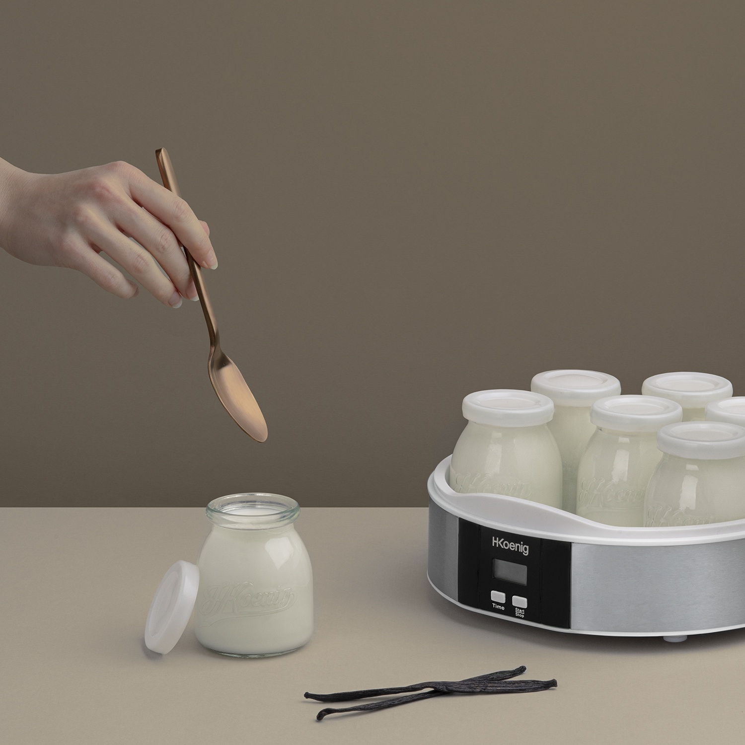 Mano tiene cucchiaio sopra vasetto yogurt; yogurtiera con vasetti, baccelli di vaniglia.