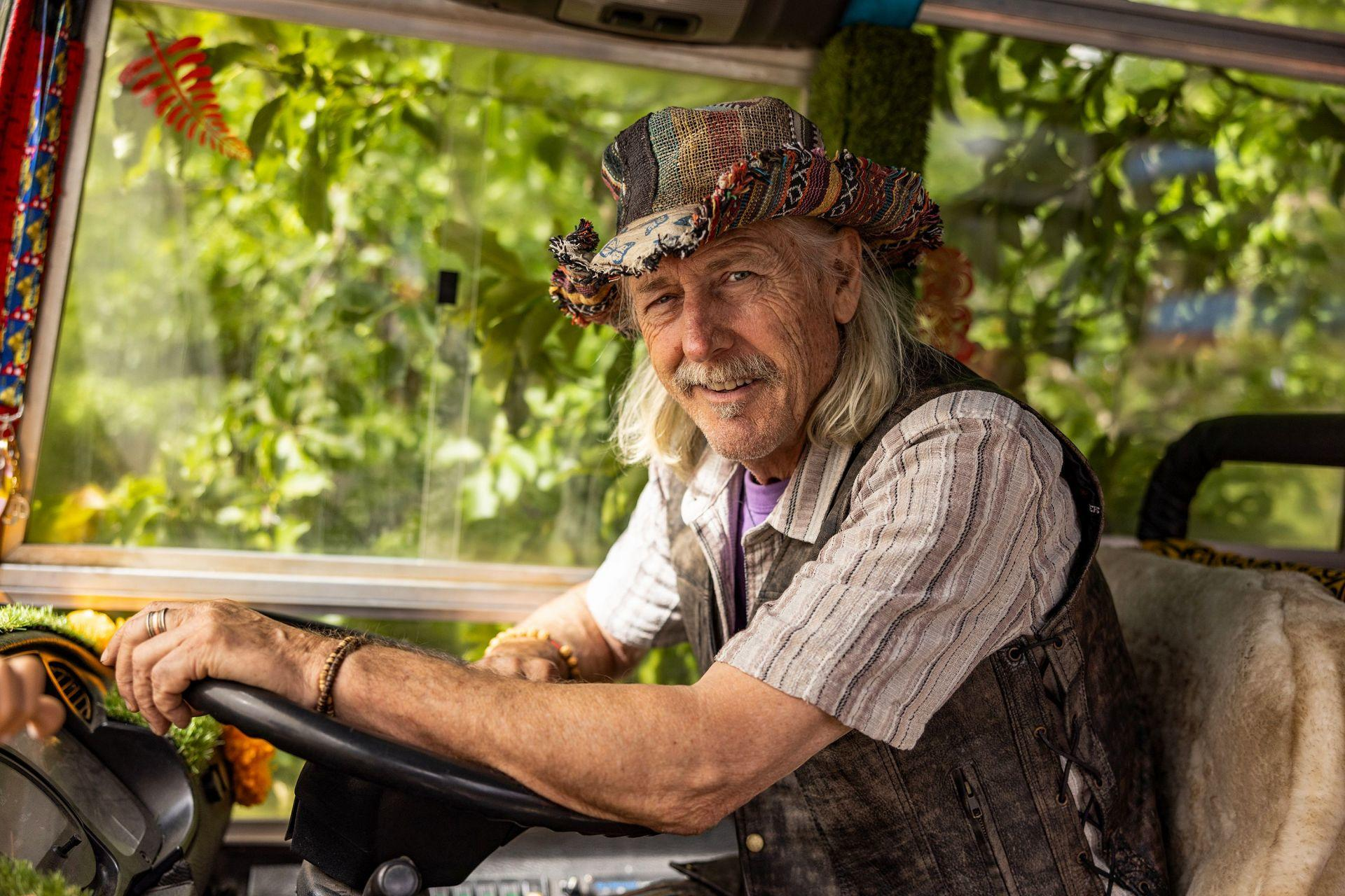 Ein Mann mit langem blondem Haar und Hut fährt einen Bus, blickt lächelnd nach vorne.