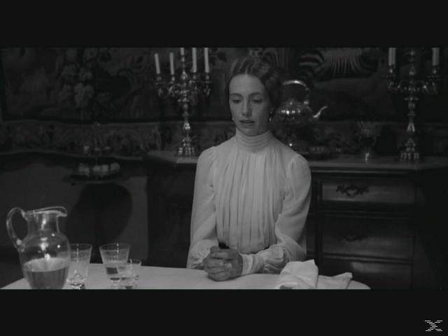 Eine Frau in einem weißen Kleid sitzt am Tisch mit einem Krug und Gläsern.
