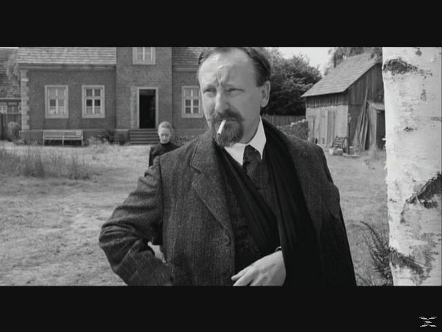 Ein Mann mit Schnurrbart raucht, eine Frau steht im Hintergrund vor einem Haus.