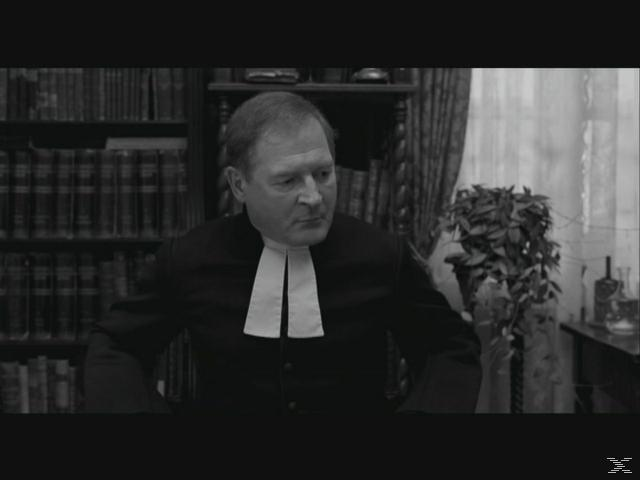 Mann in Robe vor Bücherregal und Pflanze.
