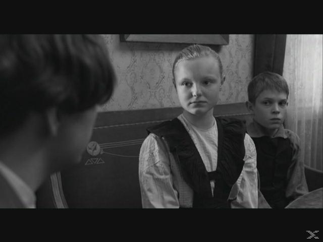 Zwei Kinder sitzen auf einer Bank. Ein Mädchen mit Zopf trägt ein dunkles Kleid. Ein Junge sitzt daneben.