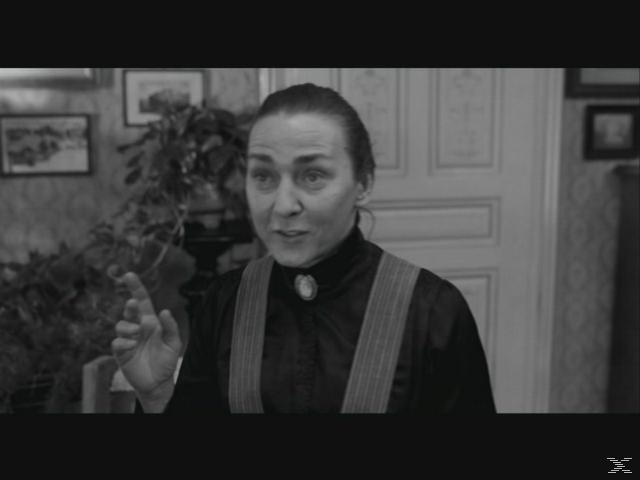 Frau in Schwarz mit Hosenträgern spricht, Hausinterieur mit Bildern.