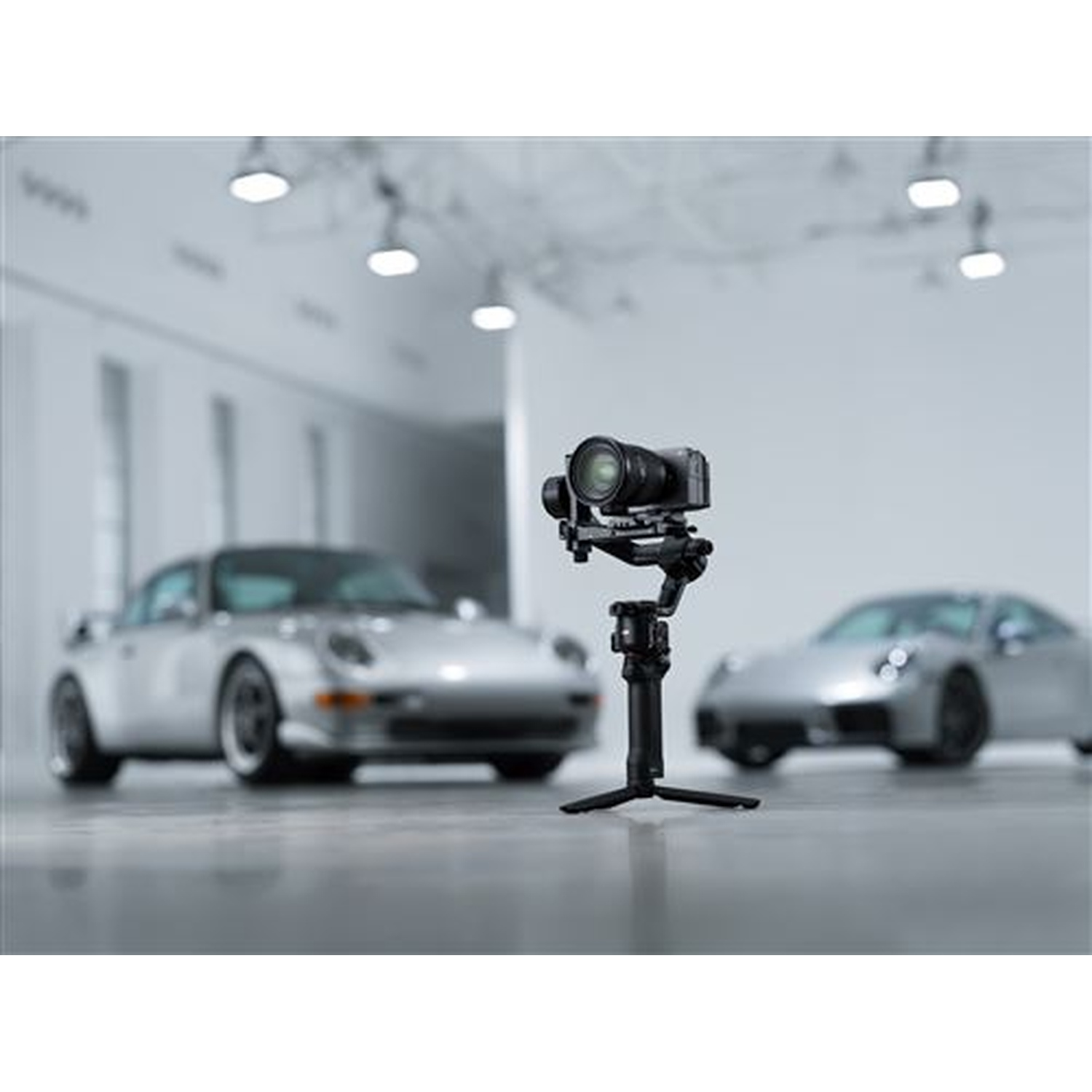 A gimbal with a camera stands before two silver sports cars in a showroom.