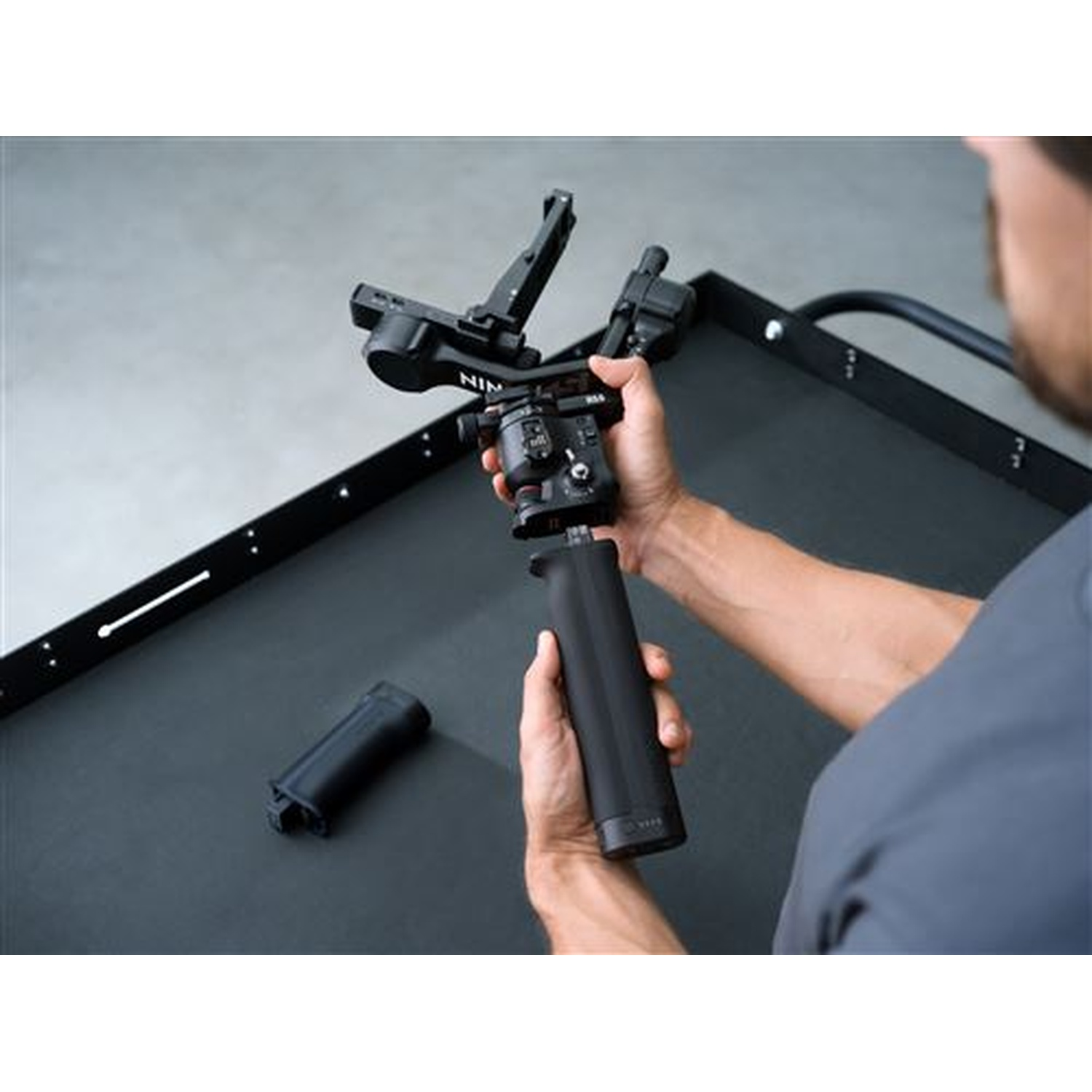 A man holds a black gimbal with a camera mount. A handle lies beside it.