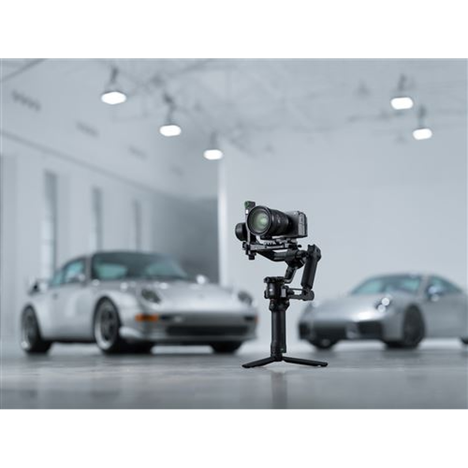 Camera gimbal in front of two silver cars in a showroom.