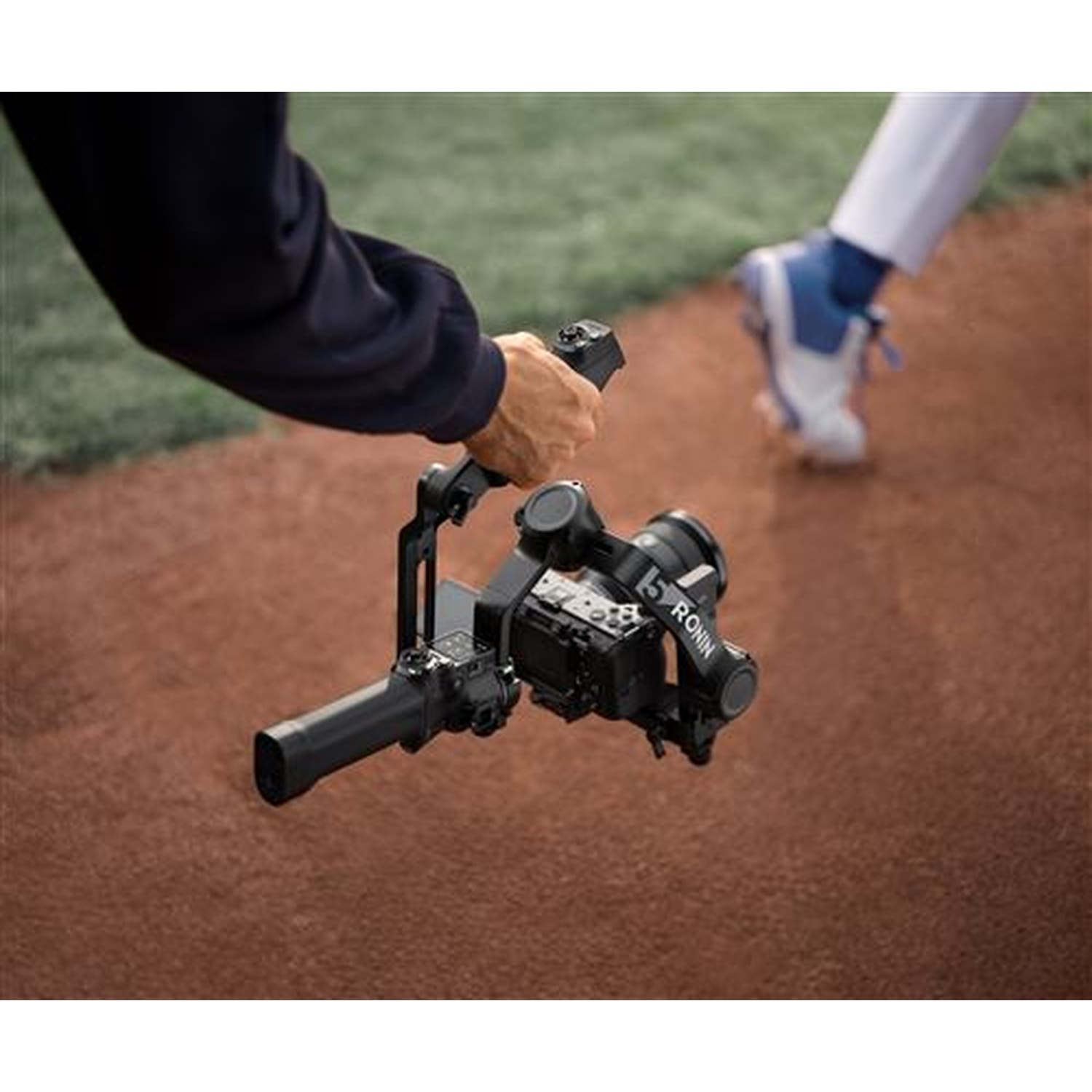 Close-up of a camera on a gimbal, held by a person in the foreground on a baseball field.