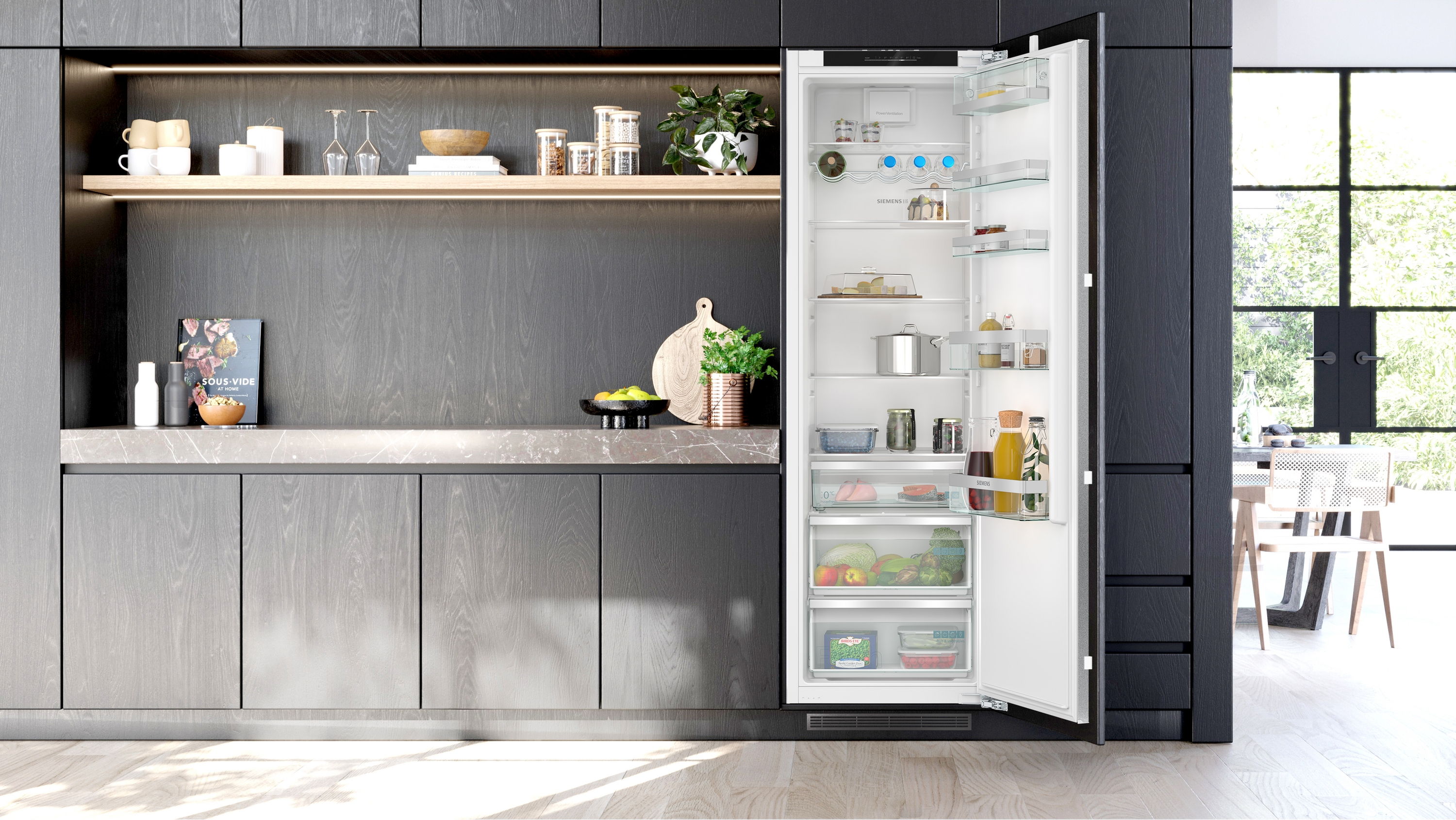 Ein offener Kühlschrank mit Lebensmitteln, daneben eine dunkle Küchenzeile mit Regalen.