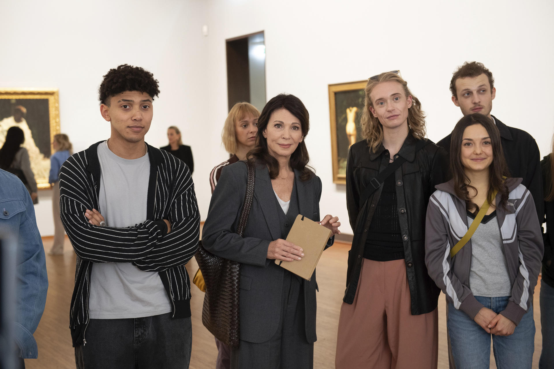 Eine Gruppe von Menschen steht in einer Galerie vor Gemälden. Eine Frau in grauem Anzug hält ein Notizbuch.