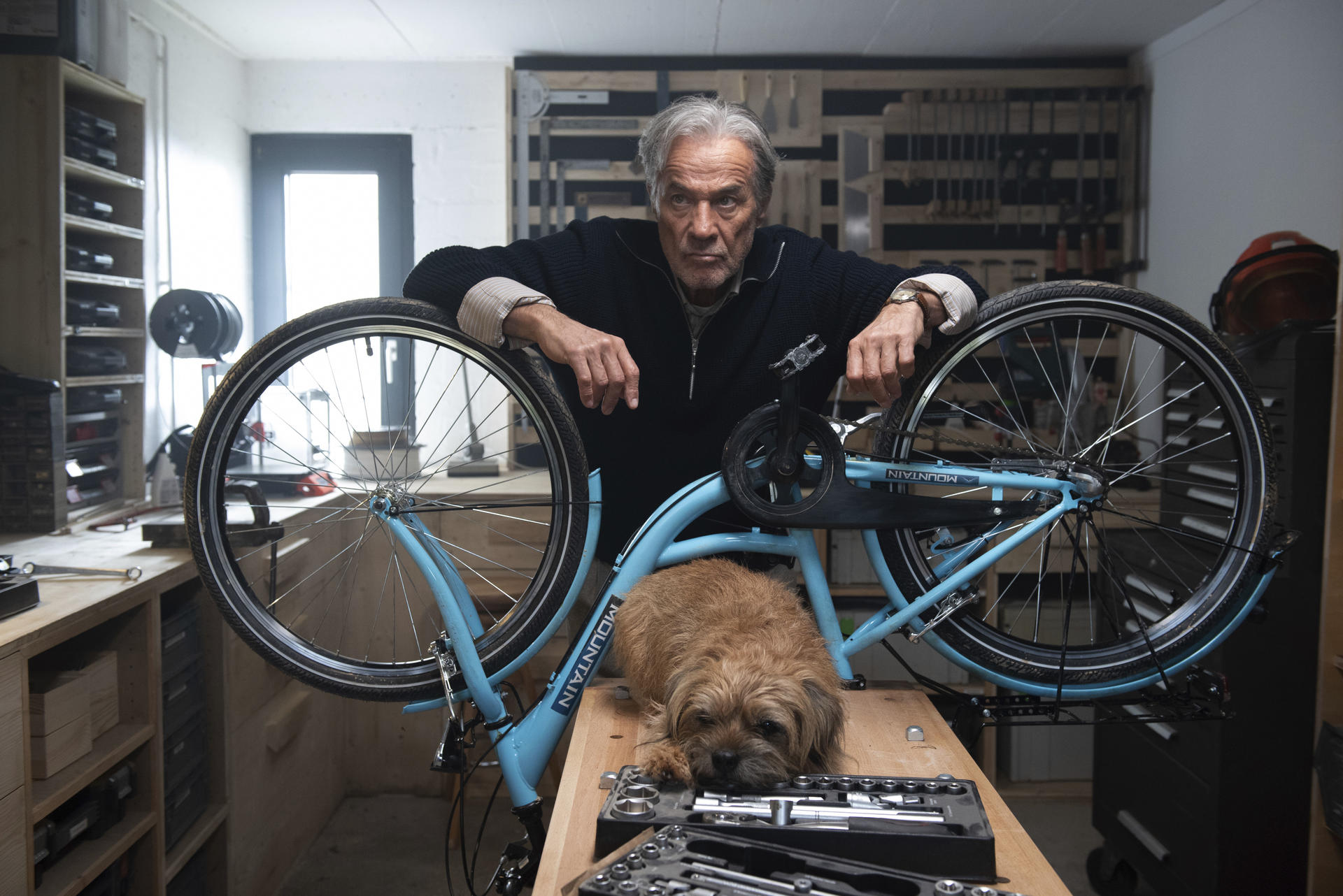 Ein Mann steht neben einem blauen Fahrrad. Ein Hund liegt davor. Werkzeuge sind auf dem Tisch.