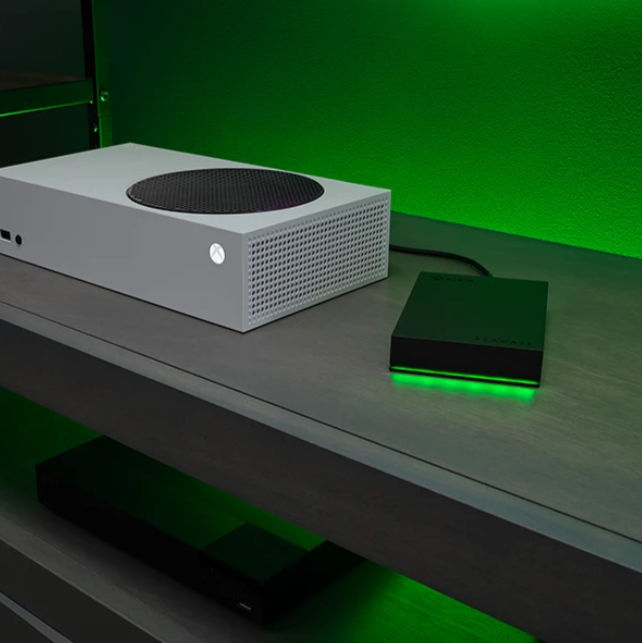 Eine weiße Xbox Konsole steht neben einer schwarzen externen Festplatte mit grüner Beleuchtung.