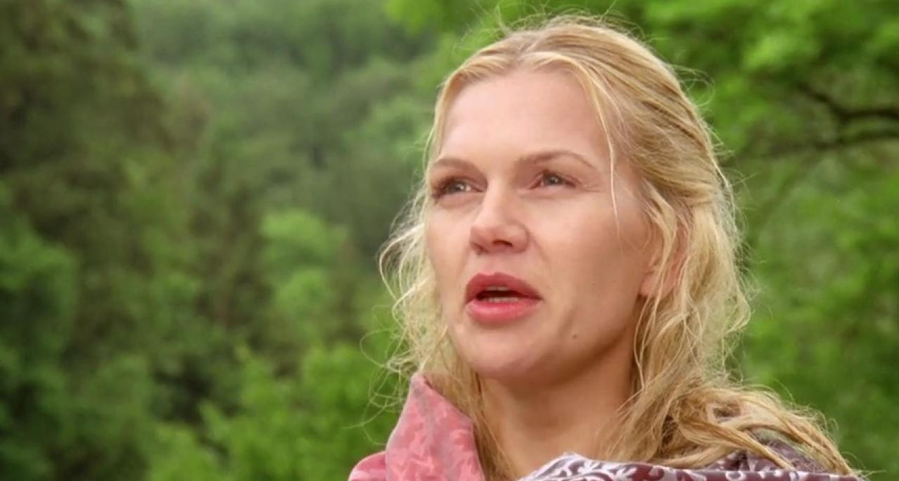 Eine blonde Frau mit roten Lippen blickt nach links, im Hintergrund grüne Bäume.