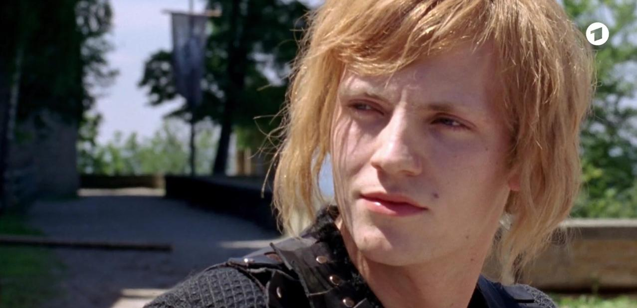 Ein junger Mann mit blonden Haaren trägt dunkle Kleidung. Er blickt nach rechts.