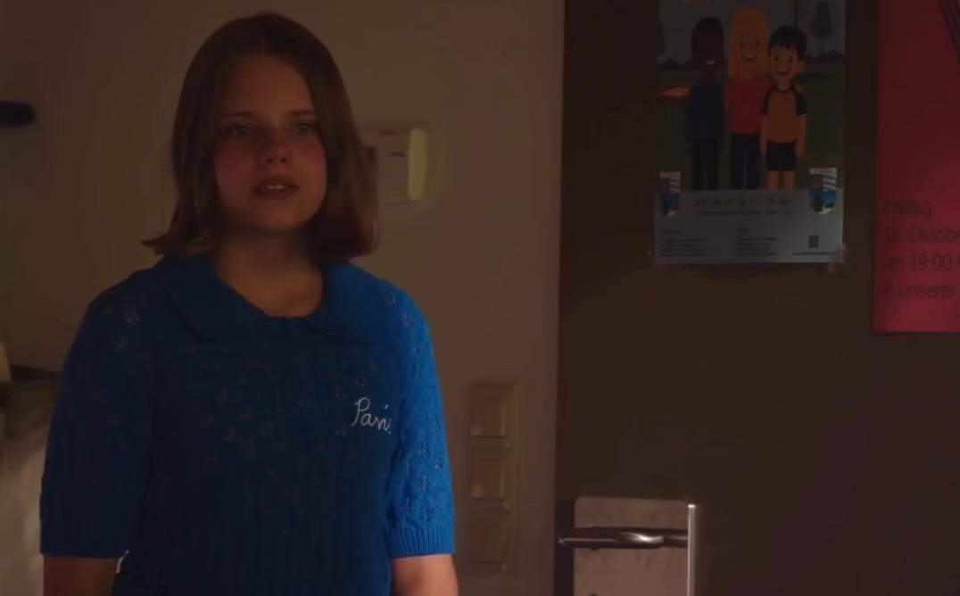 Ein junges Mädchen mit braunen Haaren in einem blauen Pullover steht vor einer Wand mit Postern.