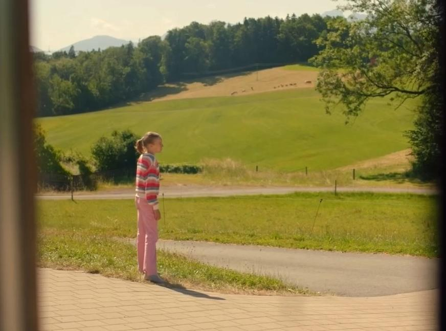Ein Mädchen steht im Freien auf einem gepflasterten Weg. Grüne Hügel im Hintergrund.