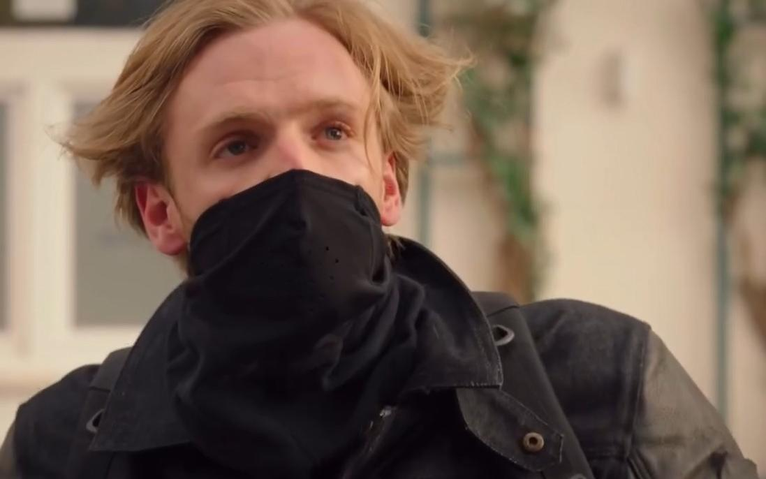 Ein Mann mit blonden Haaren trägt eine schwarze Maske und Jacke.