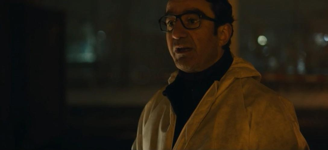 Ein Mann mit Brille trägt einen beigen Mantel und spricht.