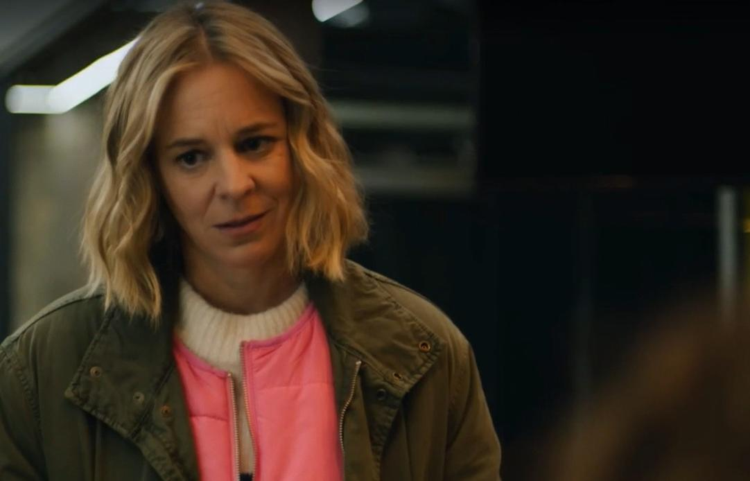 Eine Frau mit blonden Haaren trägt eine grüne Jacke über einer rosa Weste und einem weißen Pullover.